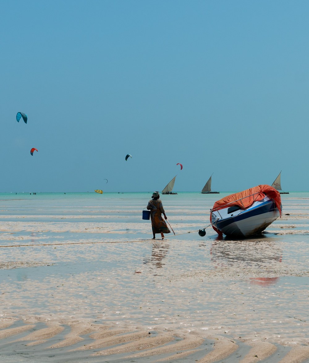 Kites fill the sky above Zanzibar's shallow coastal waters, where a person stands beside a boat.